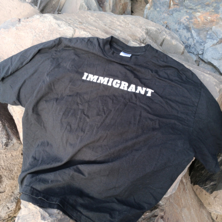 Black t-shirt with 'IMMIGRANT' printed on it, lying on rocks.